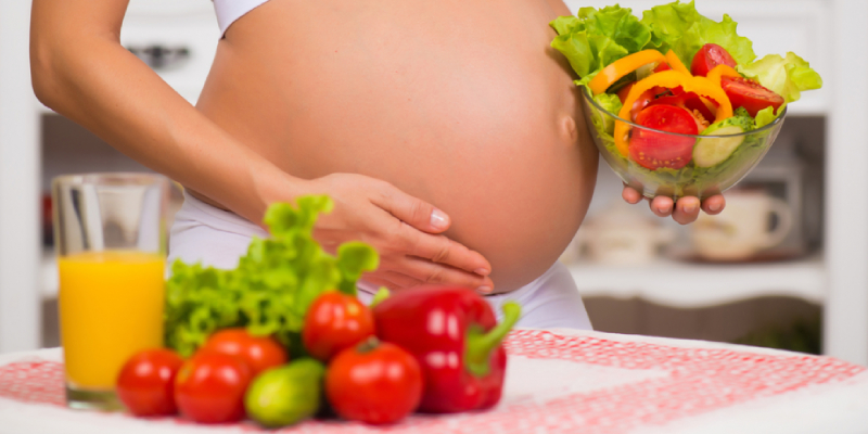 A Pregnant Woman Holds A Bowl Full Of Fresh Fruits & Vegetables.